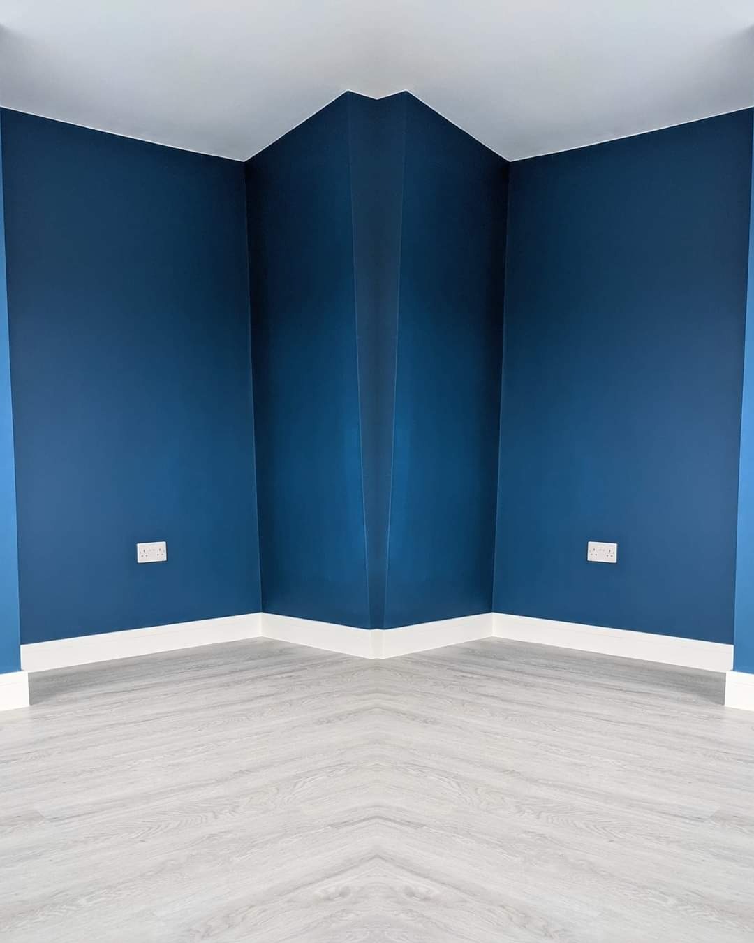Pomalowany na granatowo róg pokoju z białymi listwami przypodłogowymi i panelami podłogowymi imitującymi drewno, widoczne gniazdka elektryczne.