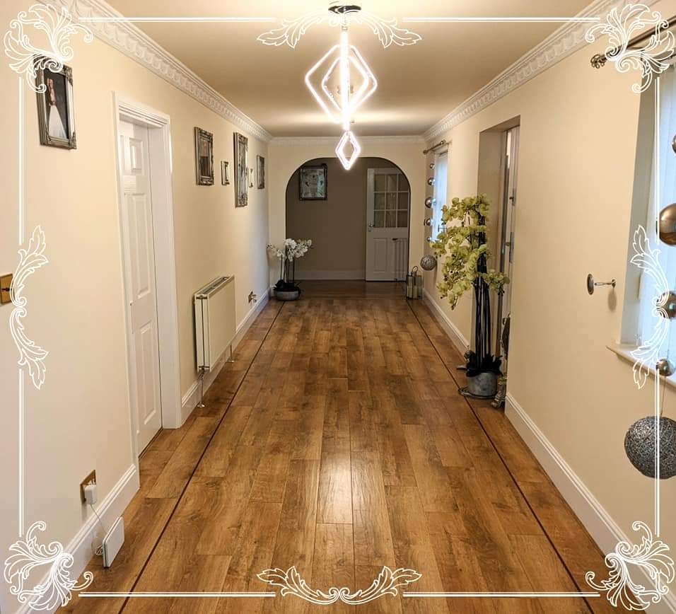 Długi korytarz z drewnianą podłogą, kremowymi ścianami, białymi drzwiami i listwami przypodłogowymi, oświetlony nowoczesną lampą wiszącą i udekorowany kwiatami w doniczkach oraz obrazami na ścianach.