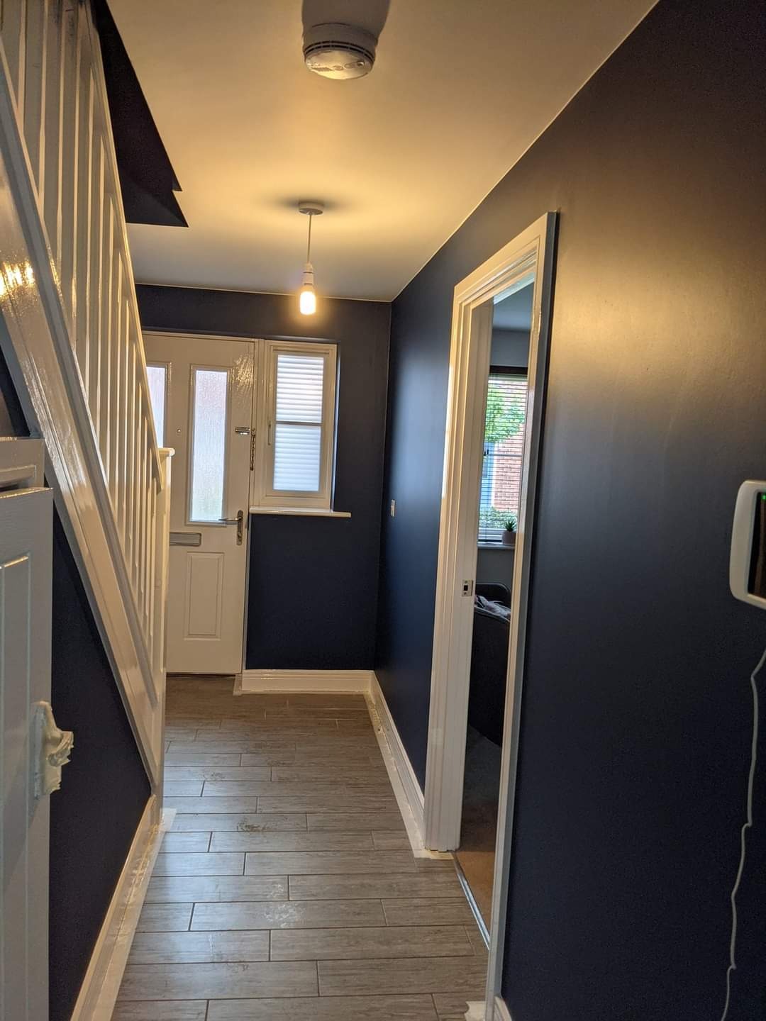 Korytarz po remoncie: ciemnoniebieskie ściany, jasne panele podłogowe imitujące drewno, białe listwy przypodłogowe i framugi drzwiowe, widoczne schody z białą balustradą.