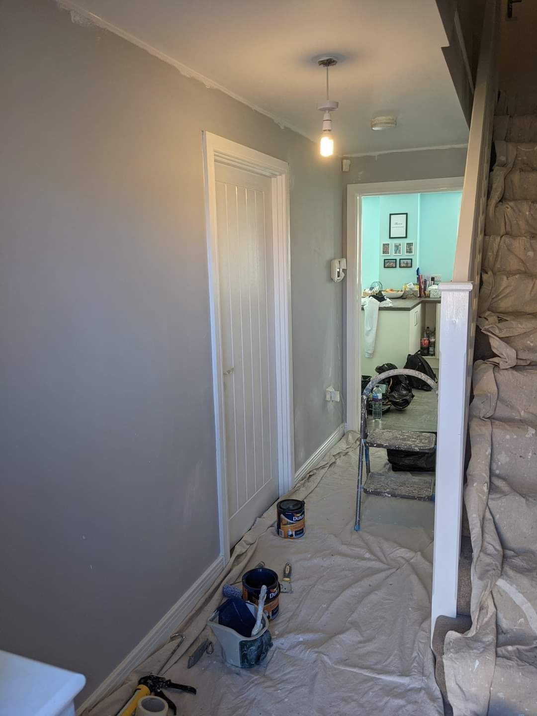 Korytarz w trakcie malowania na szaro, zabezpieczony folią malarską, widoczne puszki z farbą i drabina, świeżo pomalowane listwy przypodłogowe.