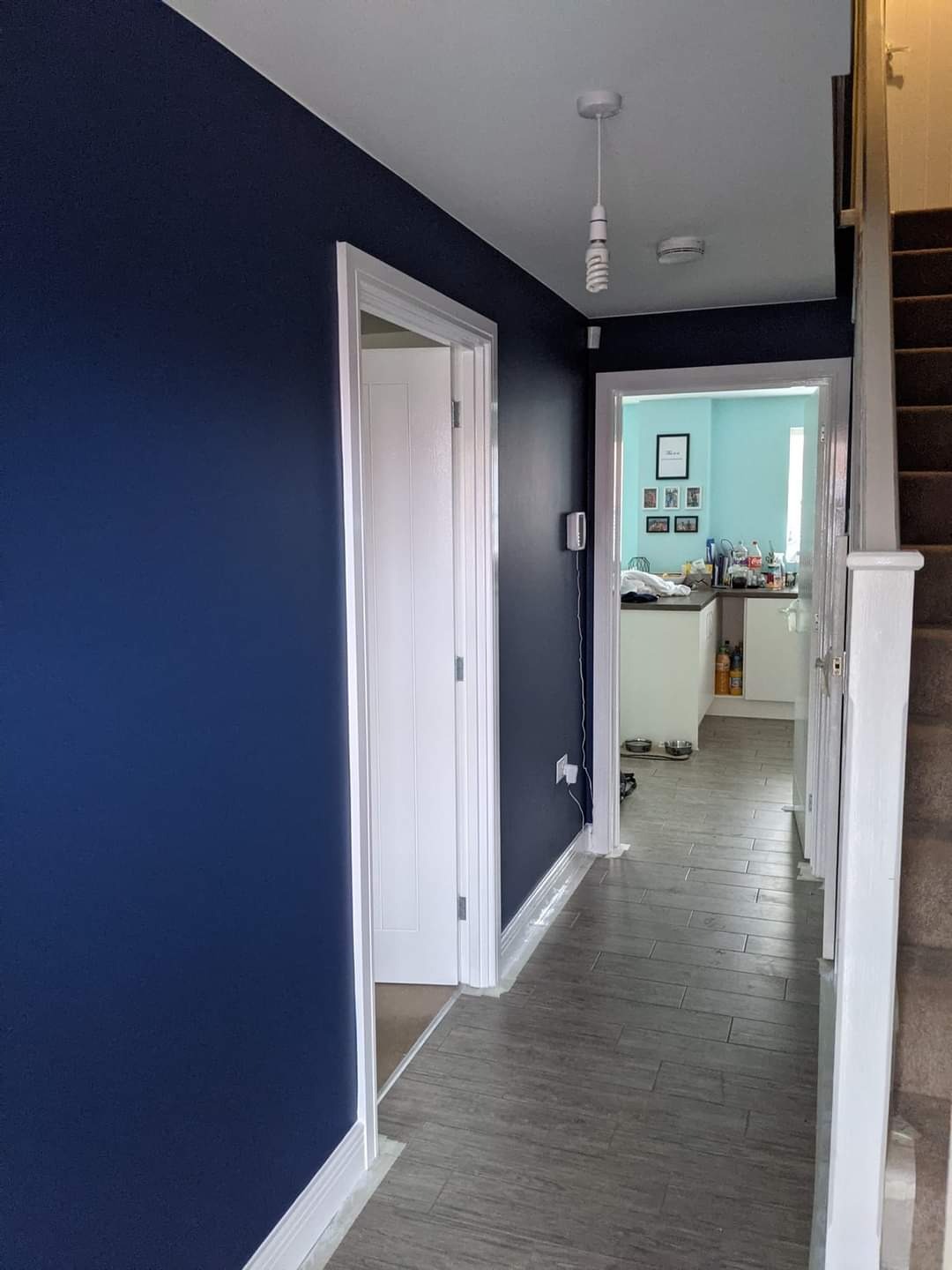Korytarz po remoncie: ściany pomalowane na granatowo, białe listwy przypodłogowe, szare panele podłogowe imitujące drewno, widok na jasną kuchnię w tle.
