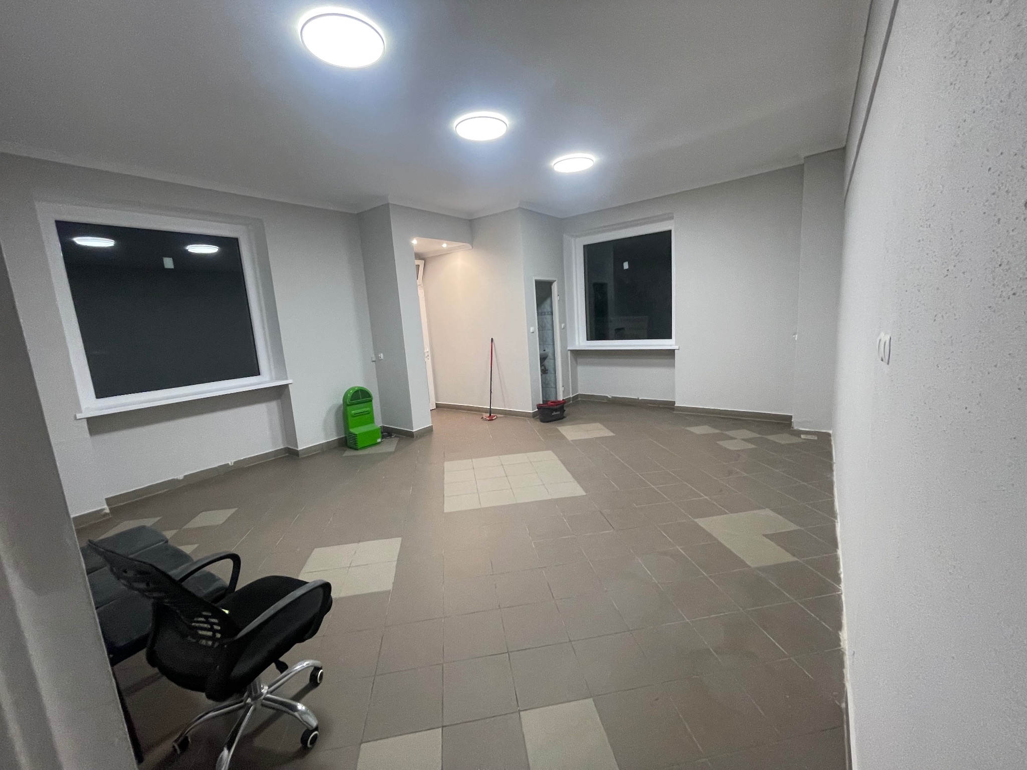 Pusta przestrzeń biurowa z szarą podłogą w kratkę, biurkiem i krzesłem na kółkach, widoczne okna, sufit z okrągłymi lampami, sprzęt do sprzątania w tle.