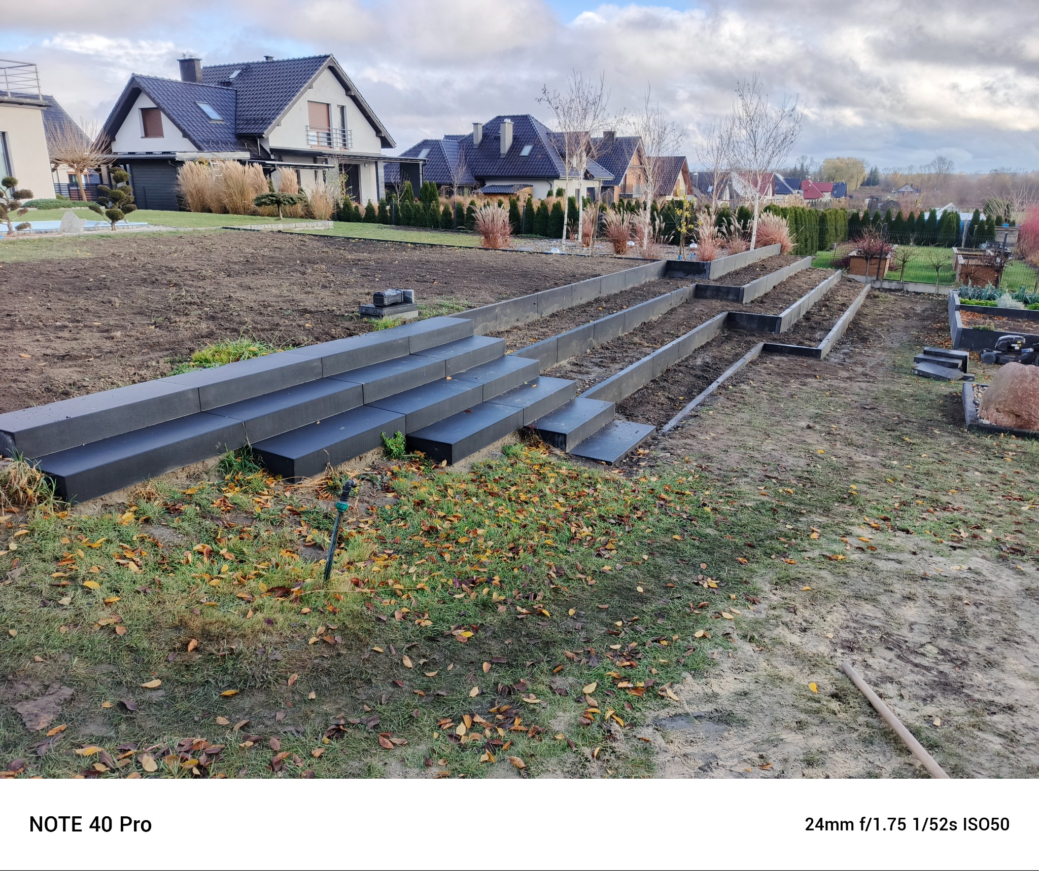 Wielopoziomowe rabaty ogrodowe z szarej kostki w trakcie budowy, widoczne przygotowane podłoże i elementy nawadniania, w tle domy jednorodzinne.