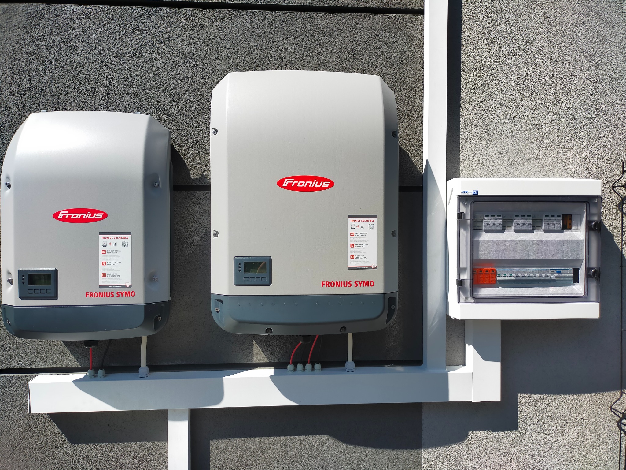 Dwa inwertery Fronius Symo zamontowane na ścianie obok skrzynki elektrycznej z bezpiecznikami, instalacja fotowoltaiczna.