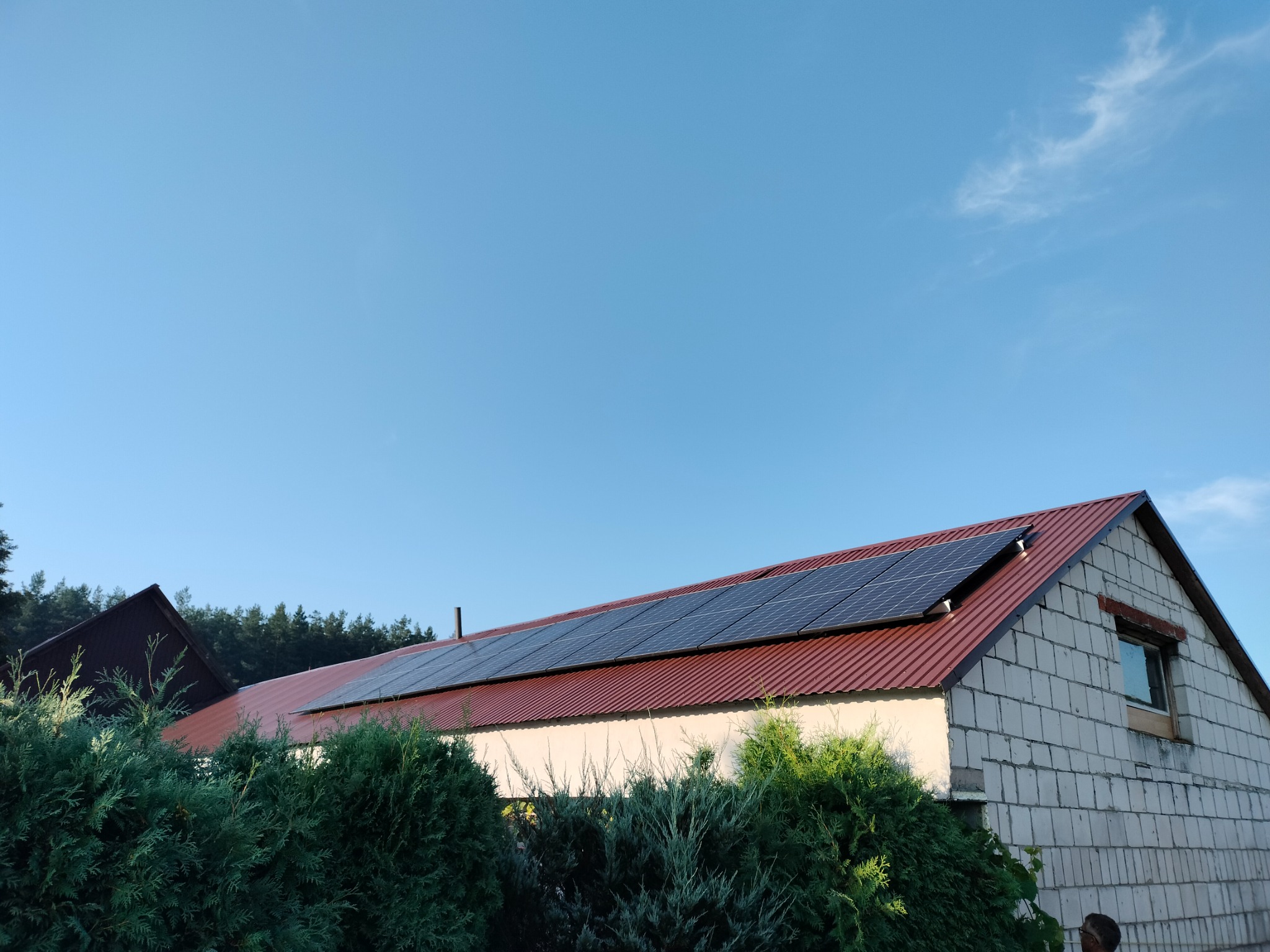 Panele słoneczne zamontowane na dachu budynku z blachy trapezowej w kolorze ceglastym, widok z dołu na tle błękitnego nieba.