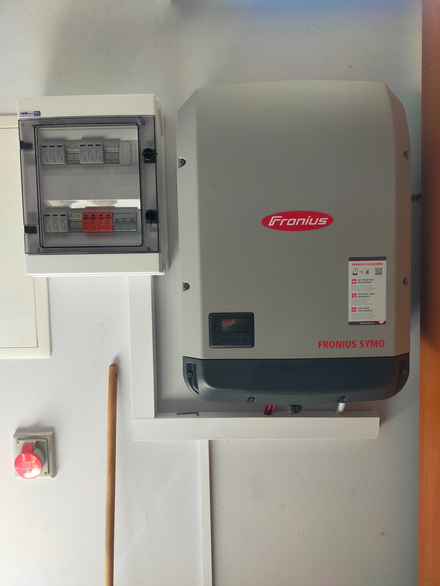 Szary falownik Fronius Symo zamontowany na ścianie obok skrzynki z bezpiecznikami i gniazda siłowego w kolorze czerwonym. Na falowniku widoczne logo Fronius i naklejka z kodem QR do monitoringu.