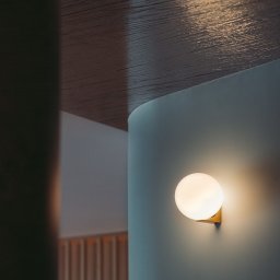 MESA STUDIO Zuzanna Stefanowicz - Kinkiet w kształcie kuli na złotej podstawie, oświetlający fragment ściany w odcieniach szarości i błękitu, z widocznym fragmentem drewnianego sufitu z widoczną fakturą i elementami dekoracyjnymi...