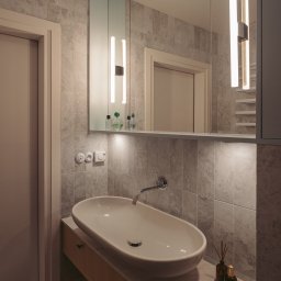 MESA STUDIO Zuzanna Stefanowicz - Nowoczesna łazienka z owalną umywalką nablatową, baterią ścienną, marmurowymi płytkami na ścianie, podświetlanym lustrem z szafką i dekoracyjnymi flakonami po prawej stronie umywalki.