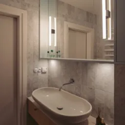 Nowoczesna łazienka z owalną umywalką nablatową, baterią ścienną, marmurowymi płytkami na ścianie, podświetlanym lustrem z szafką i dekoracyjnymi flakonami po prawej stronie umywalki.