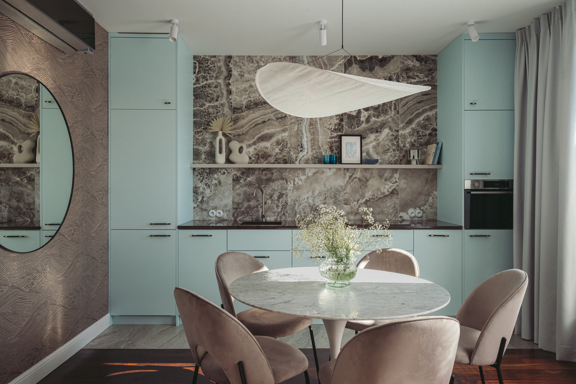 Jasna kuchnia z okrągłym stołem z marmurowym blatem i welurowymi krzesłami, dekorowana kwiatami w szklanym wazonie, oświetlona lampą w kształcie chmury.