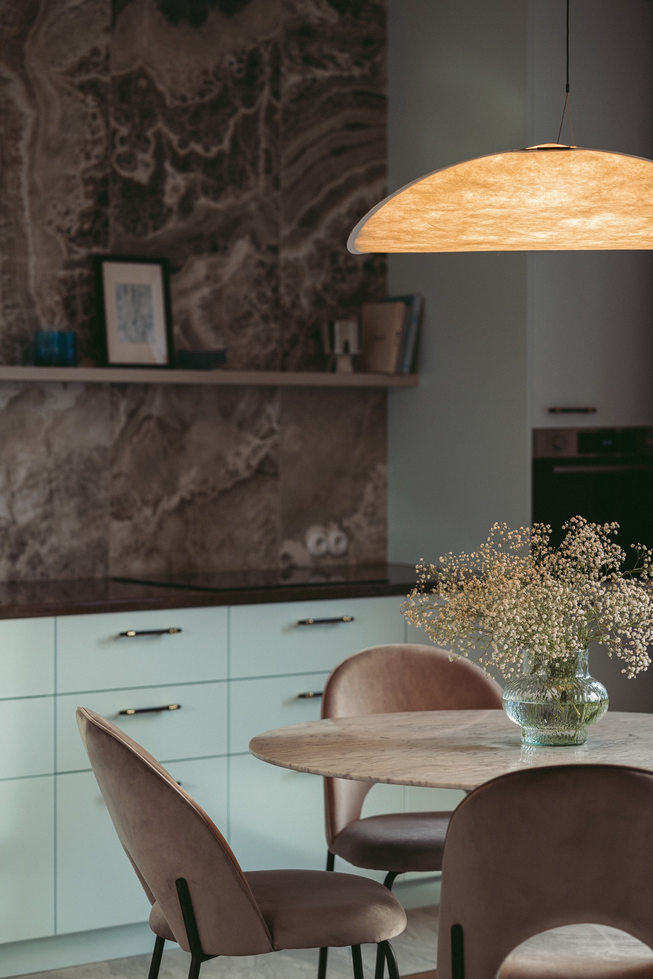 Stylowa jadalnia w Gdyni z okrągłym marmurowym stołem, welurowe krzesła w odcieniach brązu, bukiet gipsówki w szklanym wazonie i designerska lampa wisząca z abażurem z papieru ryżowego na tle...