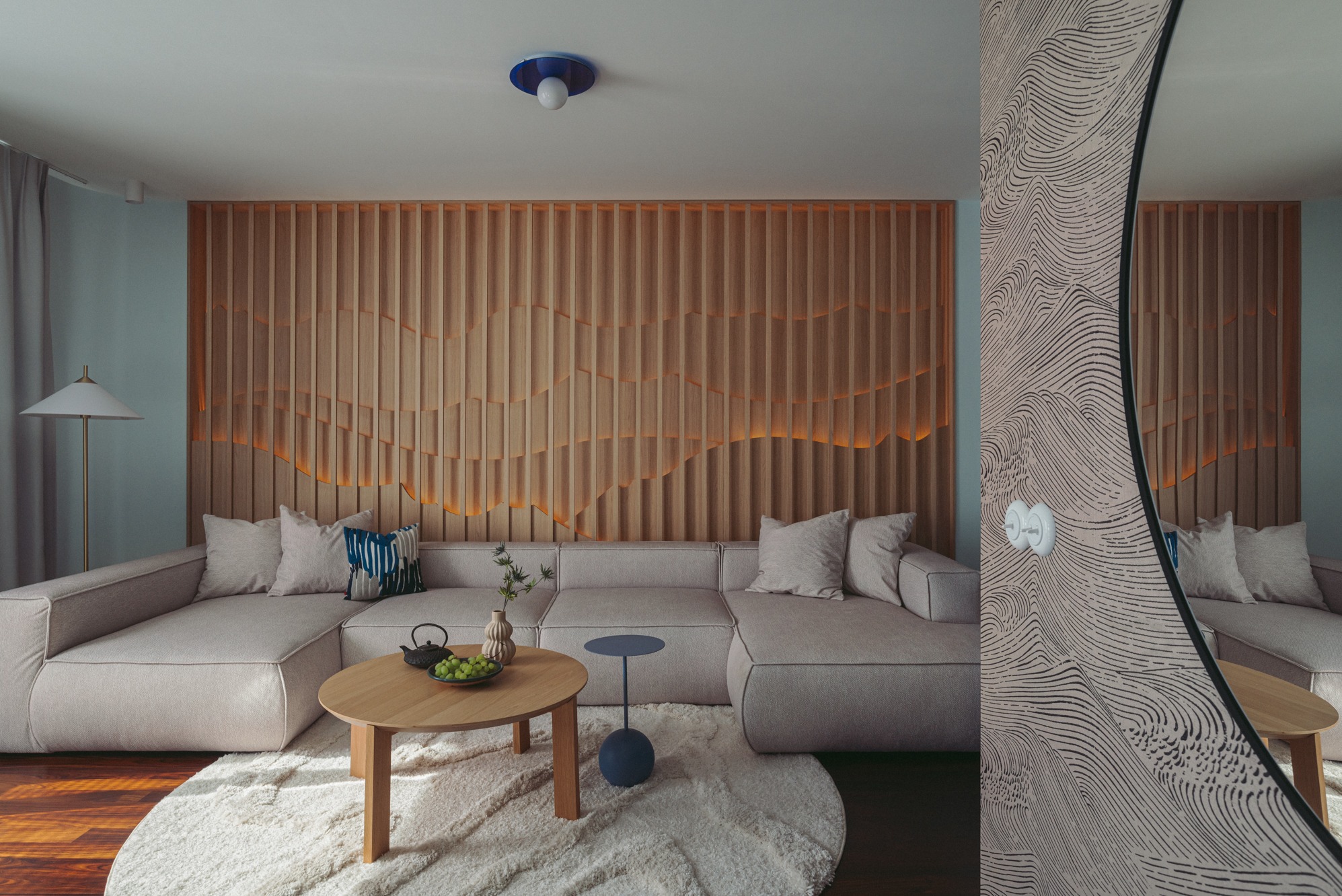 Jasny salon z beżową sofą modułową, okrągłym drewnianym stolikiem kawowym z zielonymi owocami, dekoracyjną ścianą z drewnianych listew z podświetleniem i okrągłym lustrem z wzorzystą tapetą w tle.