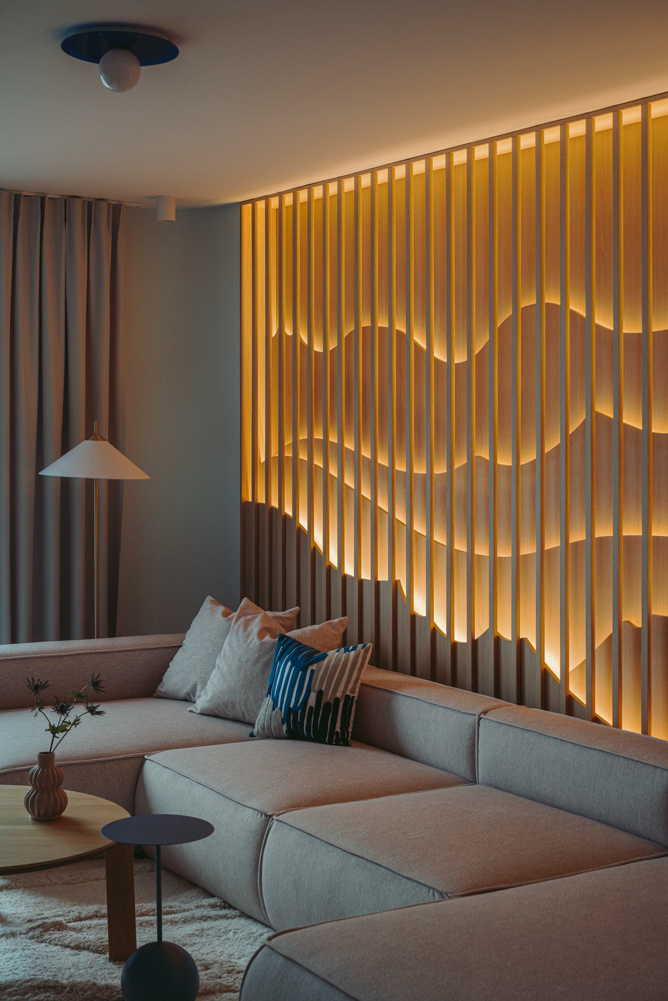 Nowoczesny salon z beżową sofą narożną, drewnianą ścianą z podświetleniem LED tworzącym falujący wzór, okrągłym stolikiem kawowym z wazonem i minimalistyczną lampą podłogową