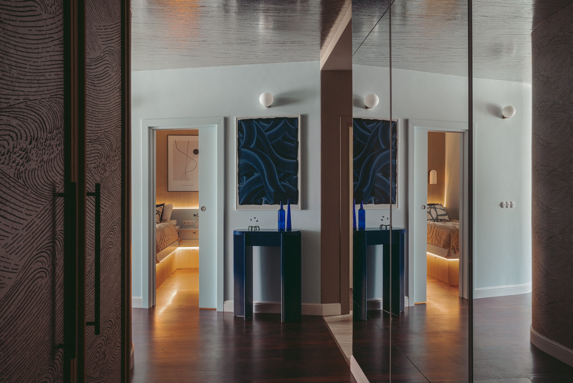 Nowoczesny korytarz z ciemną podłogą, lustrem, niebieskim stolikiem konsolowym, obrazem w ramie i widokiem na sypialnię z podświetlanym łóżkiem, uchwycony z perspektywy otwartych drzwi...