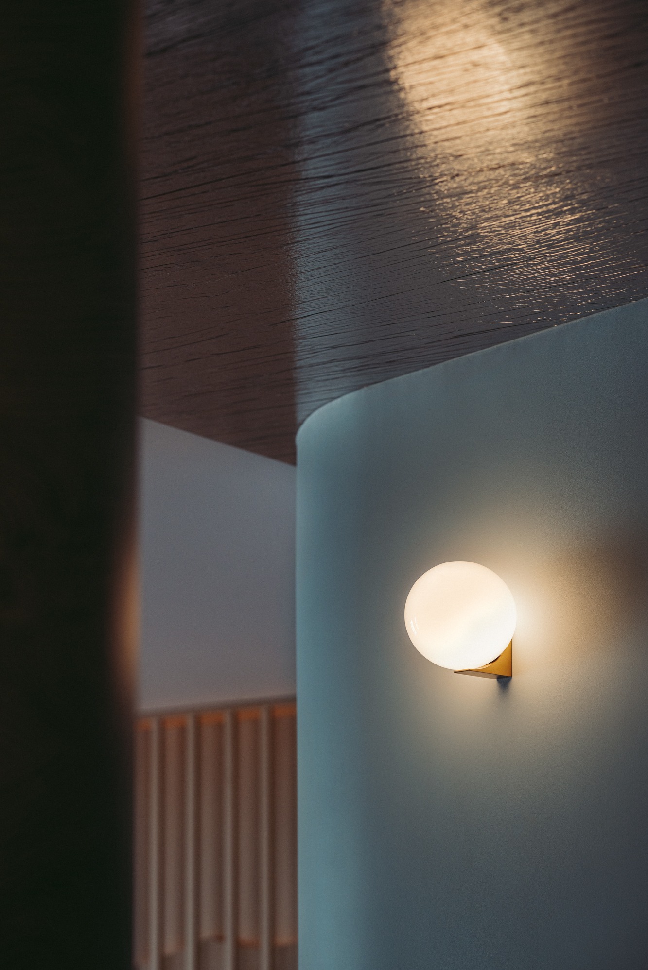 Kinkiet w kształcie kuli na złotej podstawie, oświetlający fragment ściany w odcieniach szarości i błękitu, z widocznym fragmentem drewnianego sufitu z widoczną fakturą i elementami dekoracyjnymi...