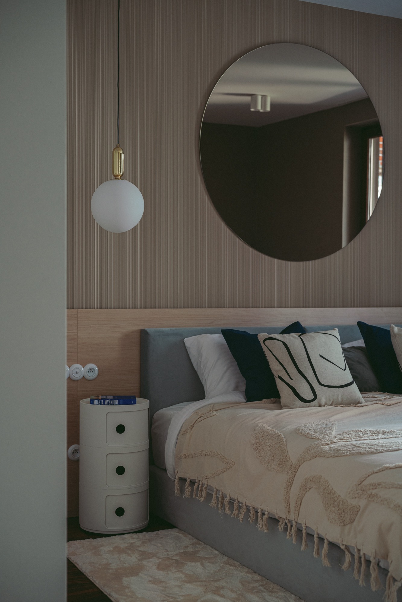 Nowoczesna sypialnia z łóżkiem w szarym obiciu, dekoracyjną narzutą z frędzlami, okrągłym lustrem na ścianie w pionowe pasy, okrągłą lampą wiszącą i stolikiem nocnym z książką 'Miasta Wysnione'.