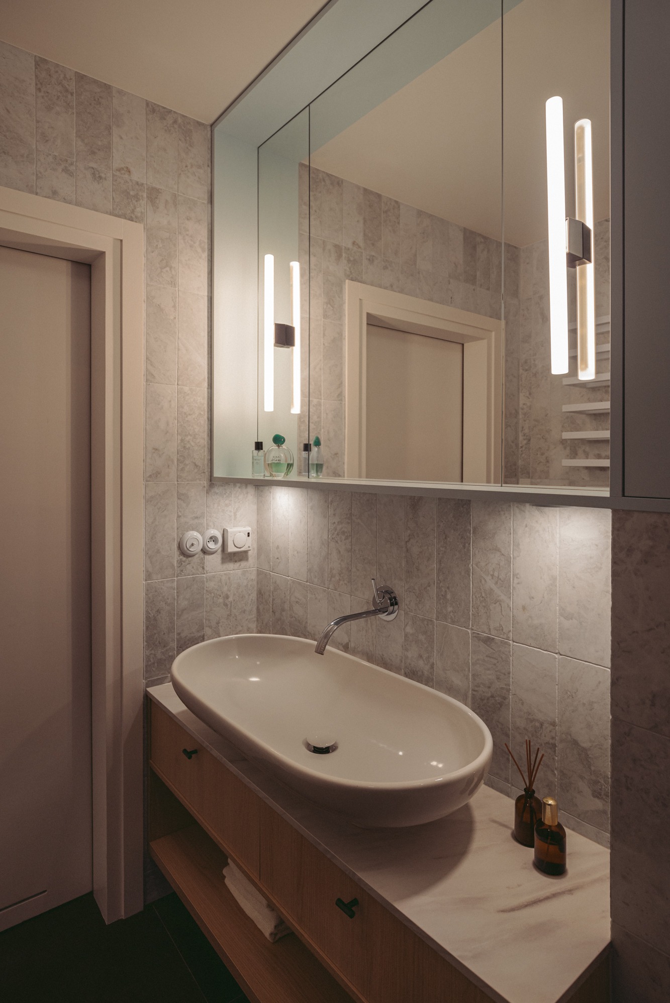 Nowoczesna łazienka z owalną umywalką nablatową, baterią ścienną, marmurowymi płytkami na ścianie, podświetlanym lustrem z szafką i dekoracyjnymi flakonami po prawej stronie umywalki.