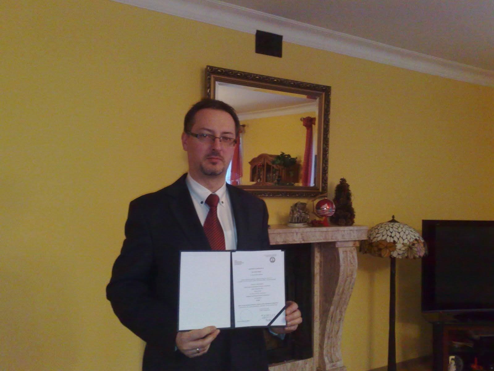 Mężczyzna w garniturze i okularach prezentuje otwarty dokument z pieczęcią na tle żółtej ściany z lustrem i kominkiem.