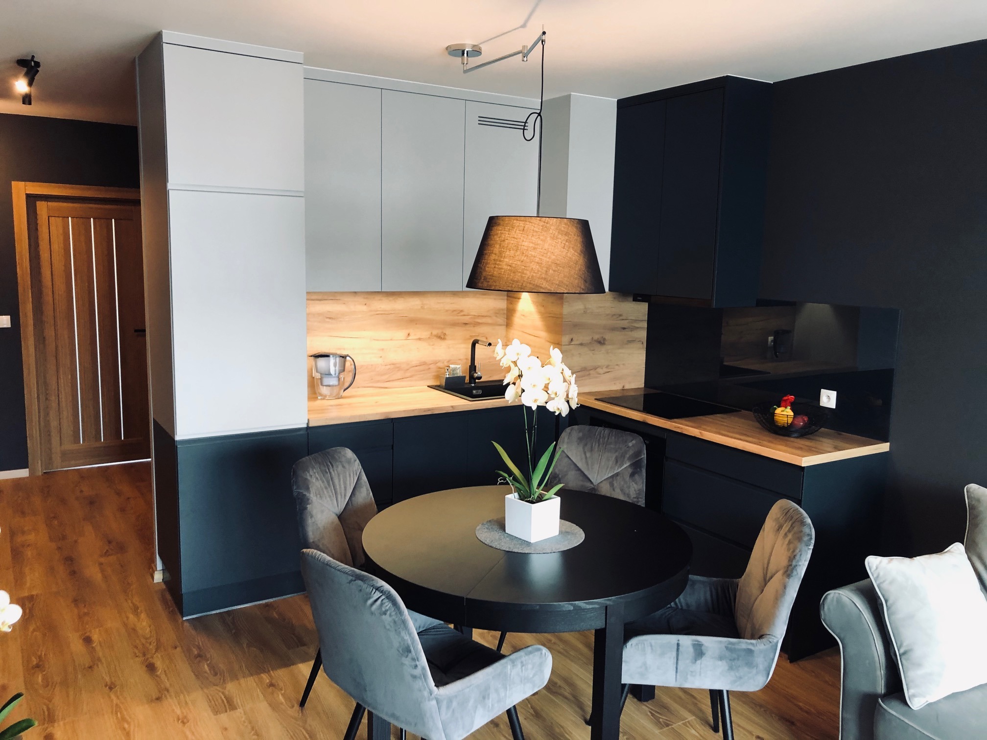 Nowoczesna kuchnia z czarnymi i szarymi szafkami, drewnianym blatem, okrągłym stołem z welurowymi krzesłami i wiszącą lampą z abażurem.