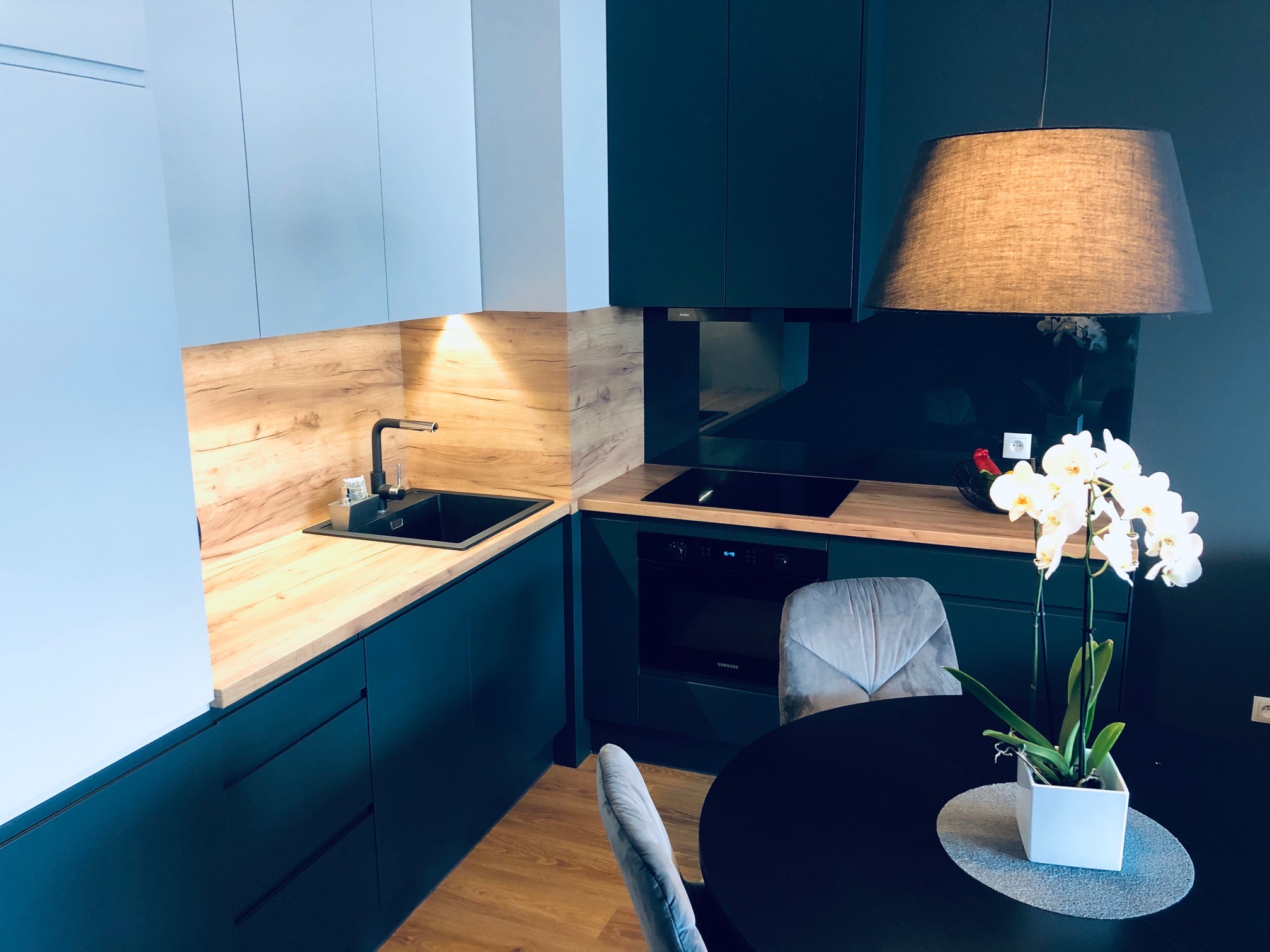 Nowoczesna kuchnia z ciemnymi szafkami dolnymi i jasnymi górnymi, drewnianym blatem, czarnym zlewem i płytą indukcyjną, oświetlona lampą z abażurem, z orchideą na stole.