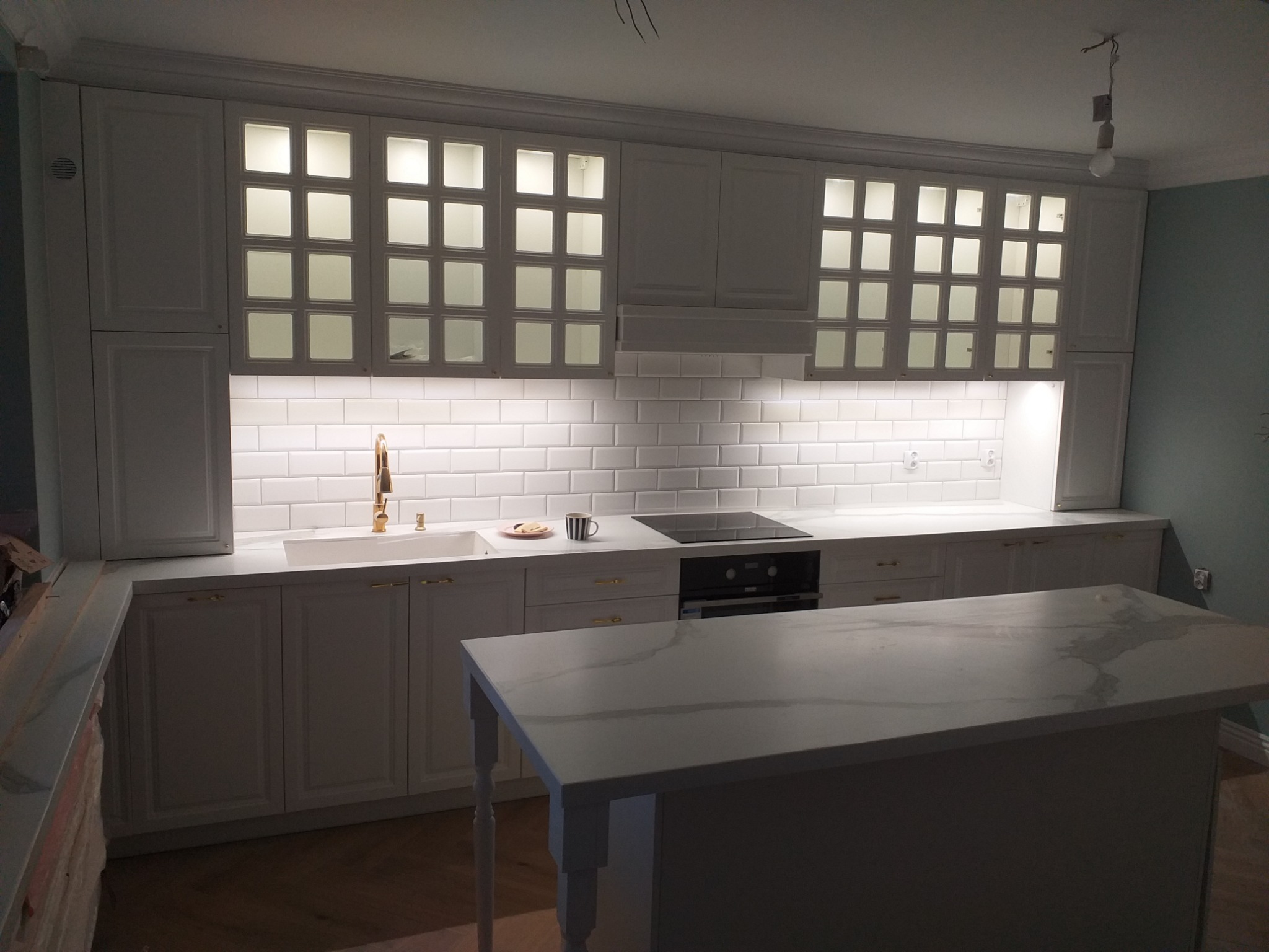 Nowoczesna kuchnia w stylu klasycznym z białymi szafkami, złotą armaturą i wyspą z marmurowym blatem. Na blacie widoczny kubek i talerzyk z ciastkami. Górne szafki przeszklone z podświetleniem.