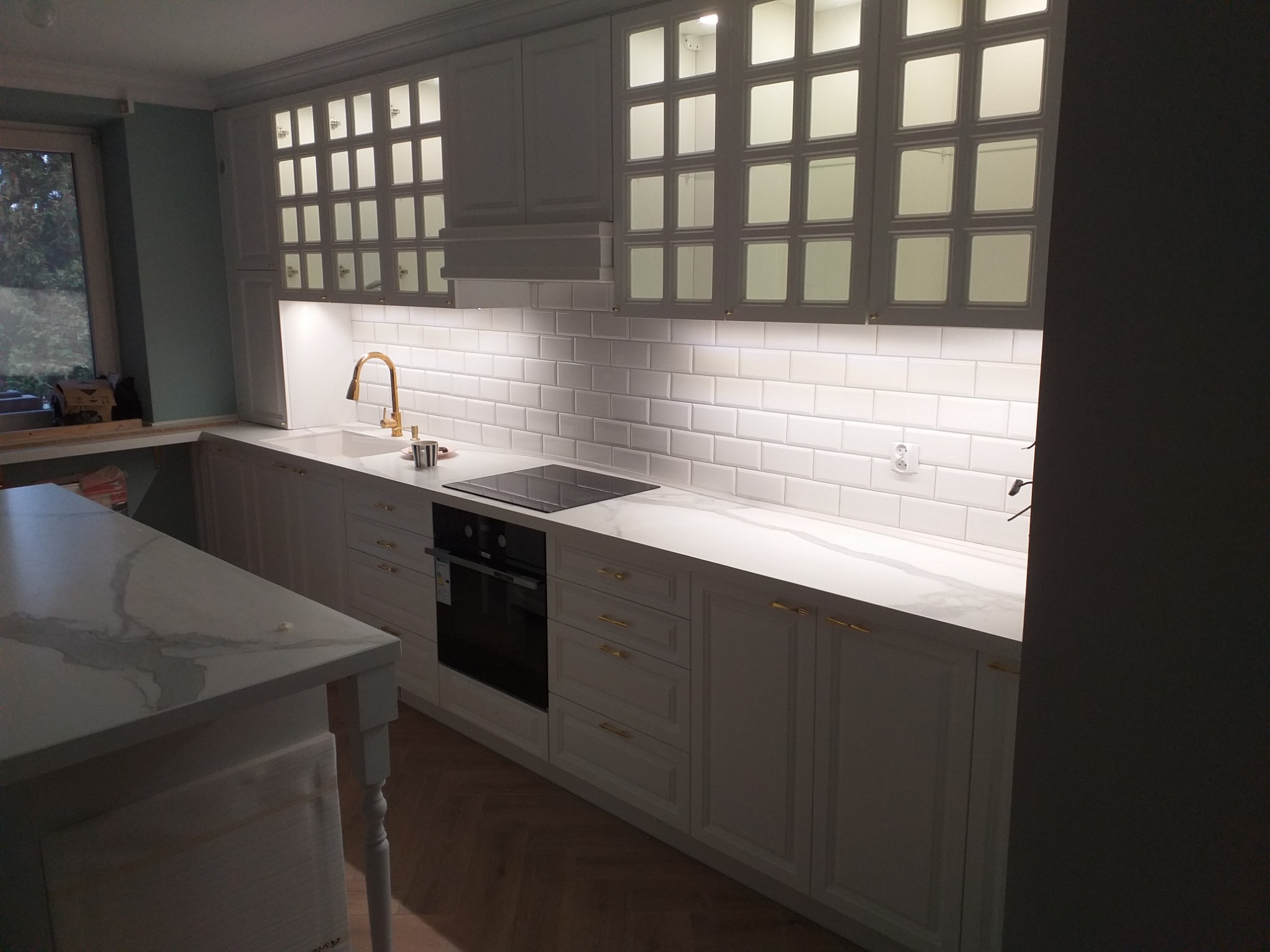 Nowoczesna kuchnia z białymi szafkami z przeszkleniami, złotą armaturą i blatem z marmurowym wzorem, oświetlona ledowym światłem podszafkowym.