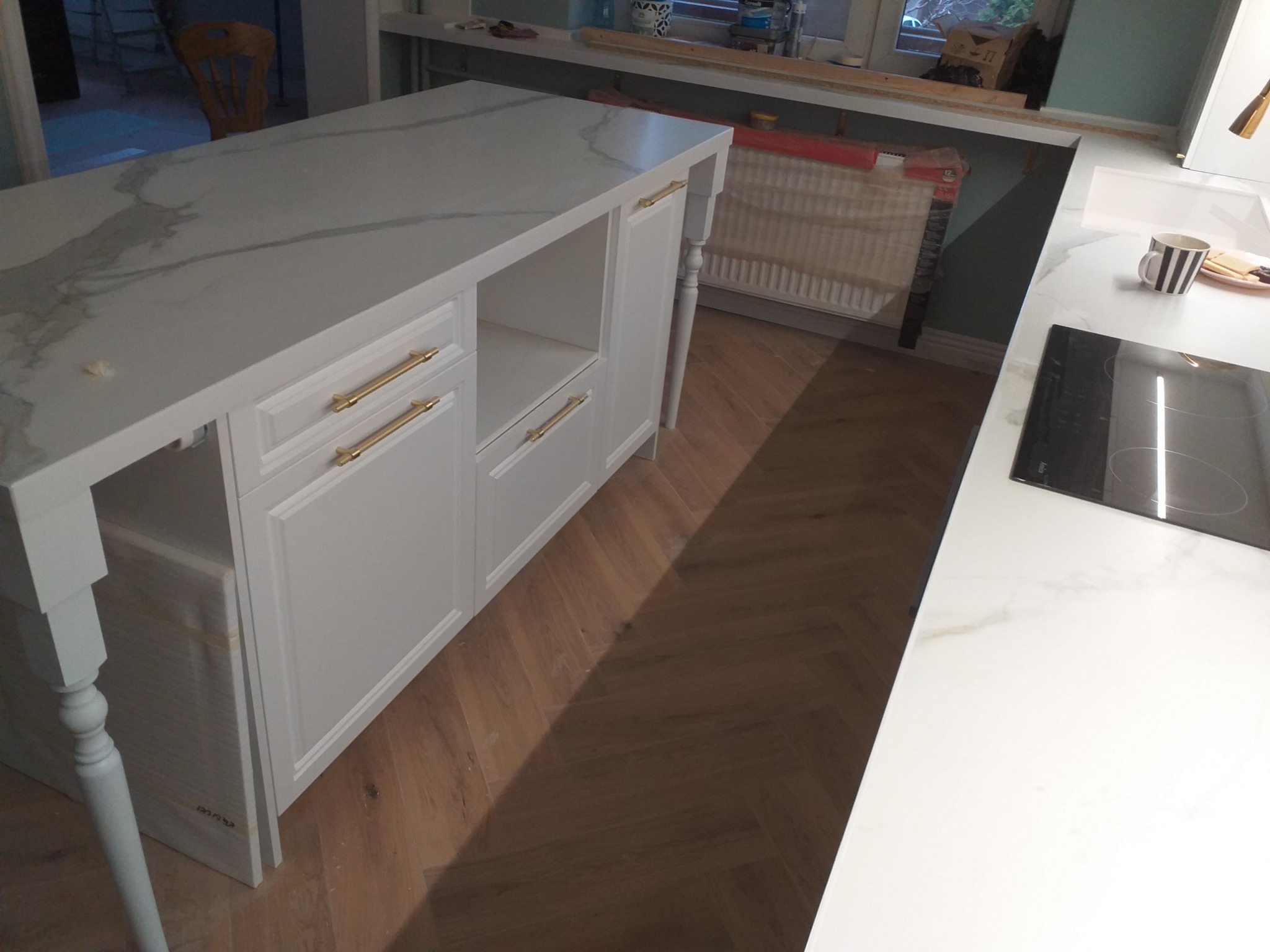 Biała wyspa kuchenna z marmurowym blatem i złotymi uchwytami, widoczna płyta indukcyjna i elementy instalacji grzewczej w tle.