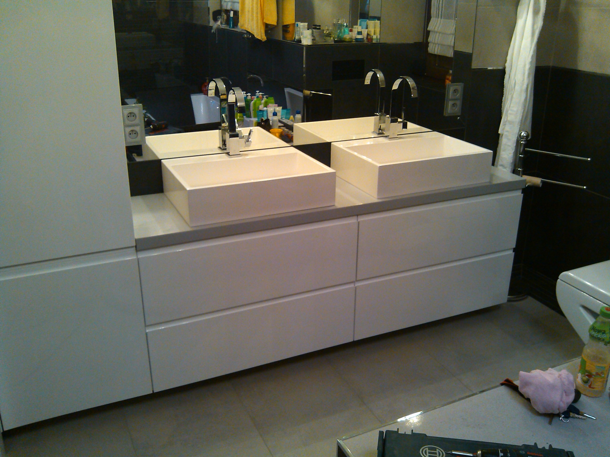 Nowoczesna łazienka z dwiema białymi, prostokątnymi umywalkami nablatowymi i chromowanymi bateriami, widoczna skrzynka z narzędziami na podłodze sugerująca prace instalacyjne.