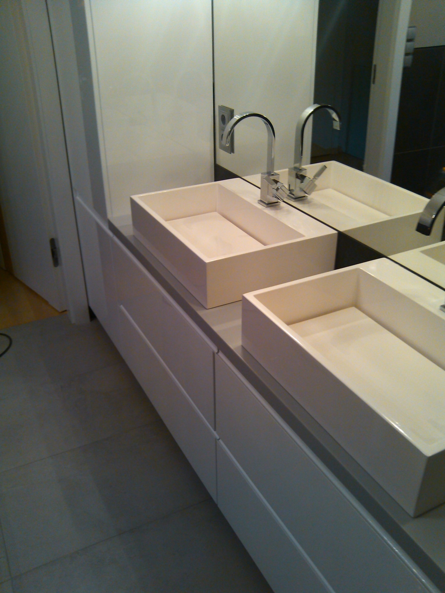 Dwie prostokątne, białe umywalki nablatowe z chromowanymi bateriami, zamontowane na białej szafce z szufladami, widoczne w lustrze.