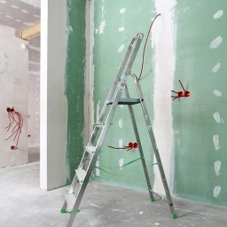 KRAWCZYK - Wnętrze w trakcie prac wykończeniowych z widoczną aluminiową drabiną, ściany częściowo pokryte płytami gipsowo-kartonowymi w kolorze zielonym, wystające kable elektryczne z puszek instalacyjnych.