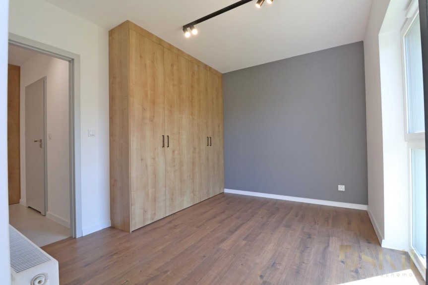 Wnętrze pokoju z drewnianą podłogą, dużą szafą z jasnego drewna i szarą ścianą, oświetlone reflektorami na szynie.