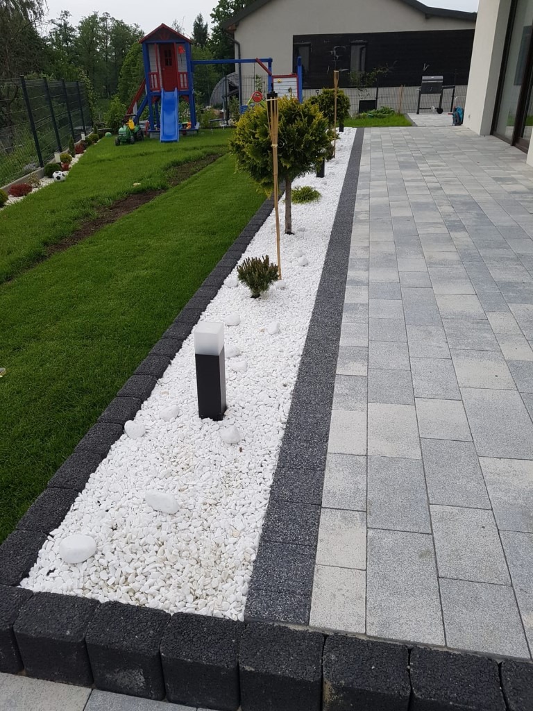 Ułożona szara kostka brukowa granicząca z rabatą wysypaną białym grysem, oddzieloną ciemnoszarym pasem, z nasadzeniami małych drzewek i nowoczesnymi lampami, w tle plac zabaw i dom.