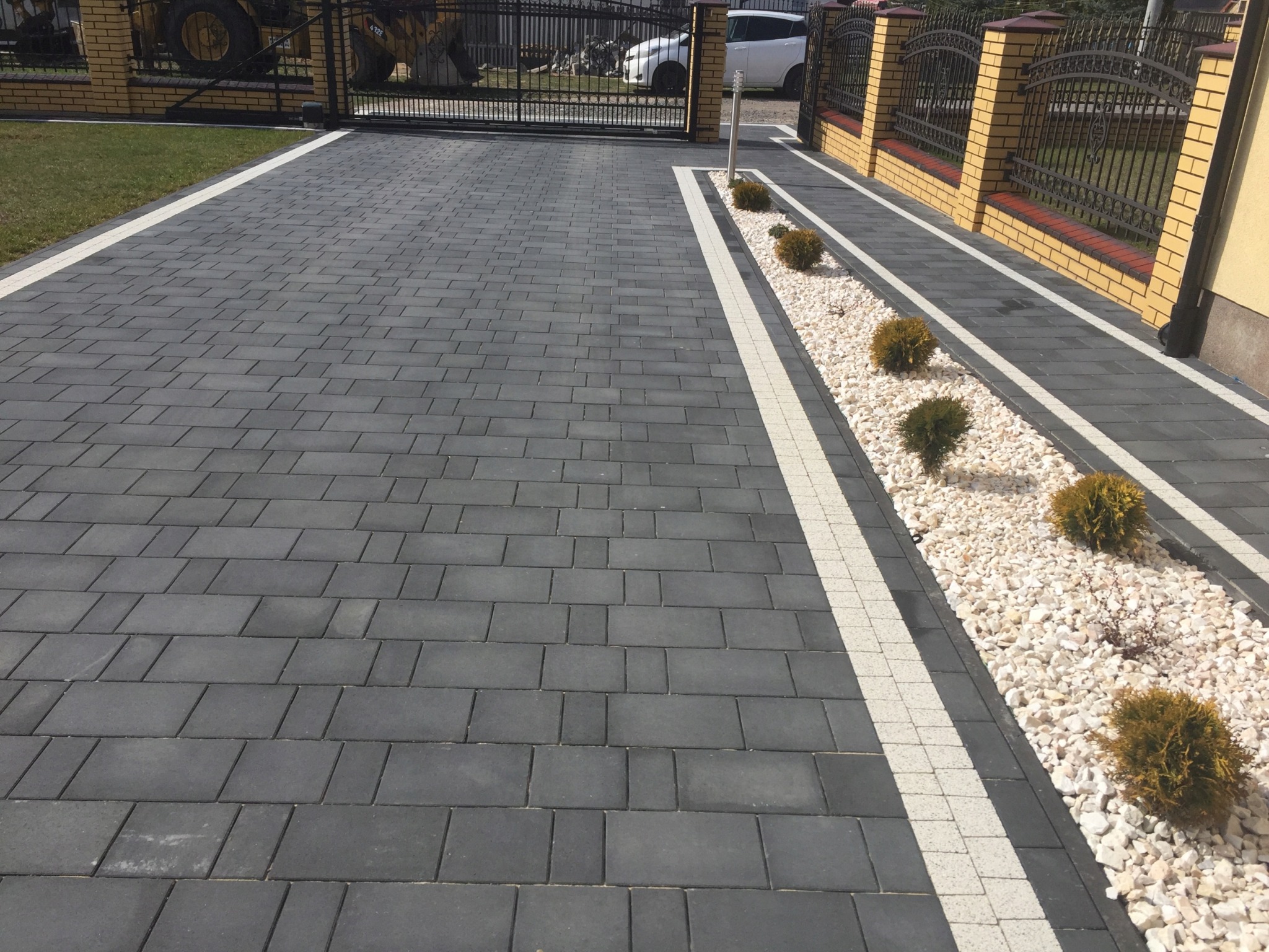 Ułożona z szarej kostki brukowej podjazd z białymi pasami oddzielającymi kostkę od rabaty z białym grysem i małymi kulistymi krzewami, w tle brama wjazdowa i ogrodzenie.