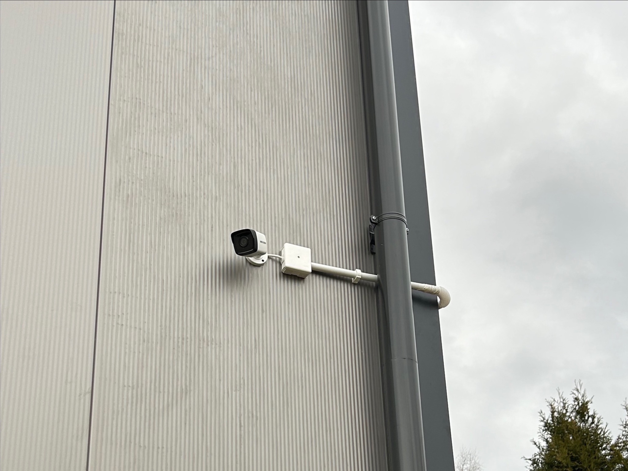Kamera monitoringu zamontowana na szarej, pionowo żłobkowanej ścianie budynku przemysłowego, obok szarej rury spustowej, widok z dołu, pochmurne niebo w tle.