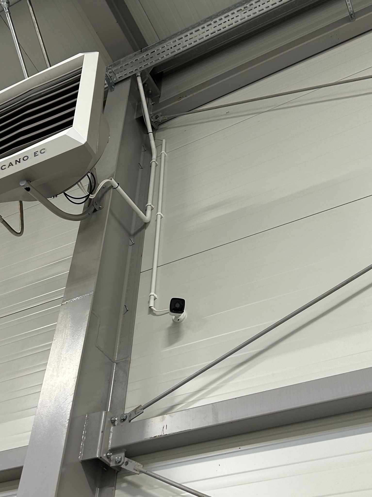 Kamera monitoringu zamontowana na ścianie hali przemysłowej, obok rur instalacyjnych i grzejnika przemysłowego Volcano EC.