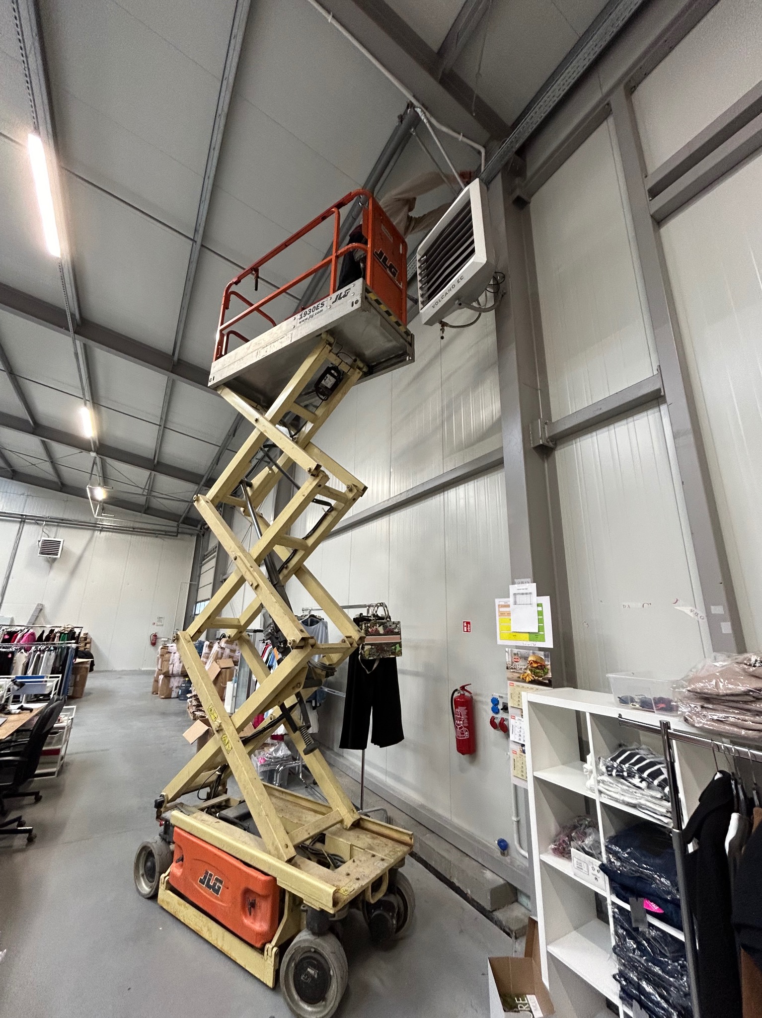 Żółty podnośnik nożycowy JLG ustawiony w magazynie z ubraniami, z widocznym nagrzewnicą Volcano EC na ścianie, sugerujący prace instalacyjne lub konserwacyjne na wysokości.