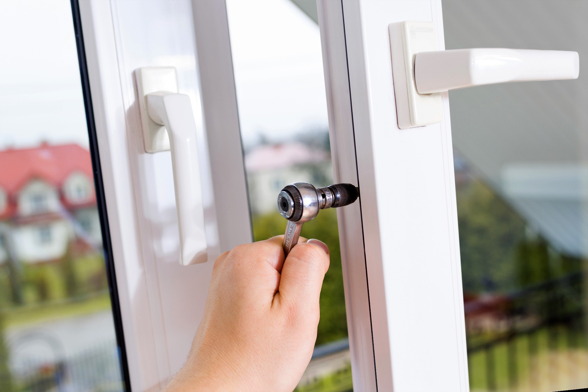 Regulacja okna PCV za pomocą klucza nasadowego; widok na dom z czerwonym dachem przez szybę.