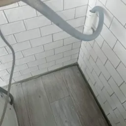 Szary wąż odpływowy pralki podłączony do białej rury na tle białych, prostokątnych płytek ściennych i szarej podłogi imitującej drewno.