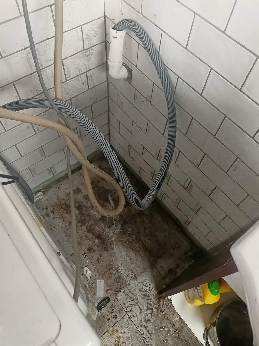 Brudna podłoga w łazience przed sprzątaniem, widoczne rury odpływowe i białe płytki na ścianach.