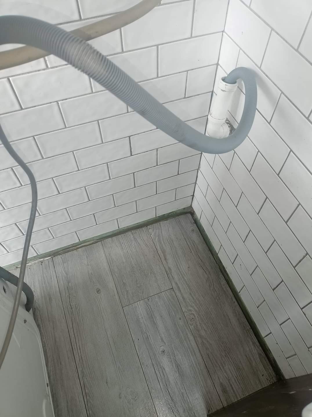 Szary wąż odpływowy pralki podłączony do białej rury na tle białych, prostokątnych płytek ściennych i szarej podłogi imitującej drewno.