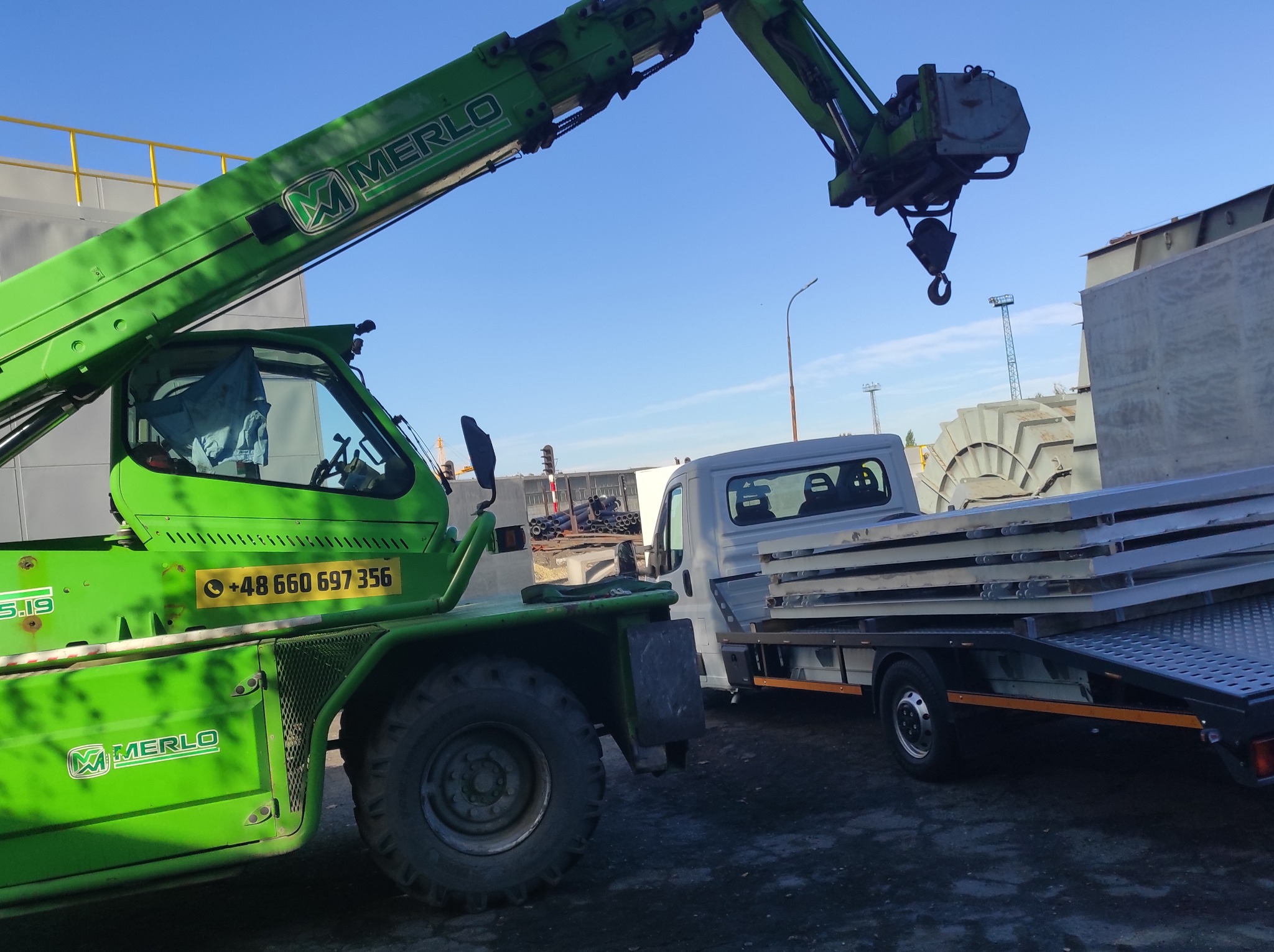 Zielony wózek widłowy teleskopowy marki Merlo podnosi za pomocą haka pakiet metalowych płyt na platformę białego samochodu dostawczego, na tle błękitnego nieba i konstrukcji przemysłowych.