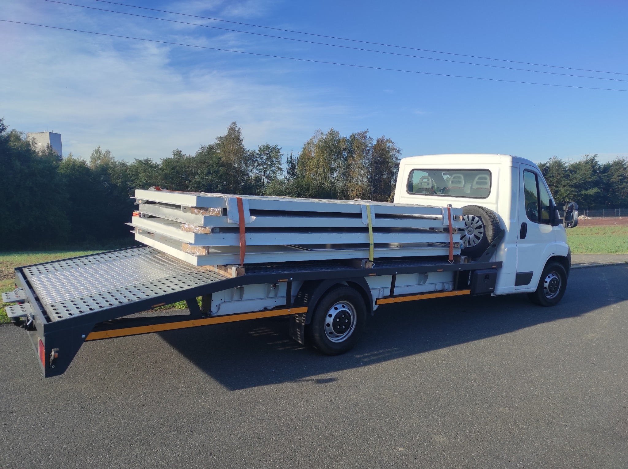 Biała laweta z opuszczoną platformą transportuje zabezpieczone pasami metalowe elementy konstrukcyjne na tle błękitnego nieba i zieleni.