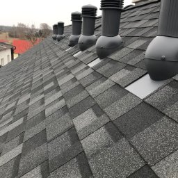 Michał Wieczorek DOMY DREWNIANE ,SZKIELETOWE. OBSŁUGA KOMPLEKSOWA - Szary dach pokryty gontem bitumicznym z zamontowanymi odpowietrznikami kominkowymi w rzędzie, widok z bliska pod kątem.