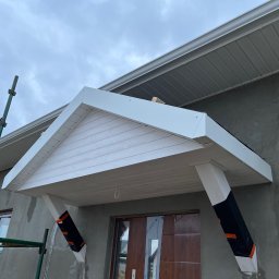 Michał Wieczorek DOMY DREWNIANE ,SZKIELETOWE. OBSŁUGA KOMPLEKSOWA - Nowo wybudowany, biały, zadaszony ganek z drewnianymi kolumnami zabezpieczonymi folią, na tle surowej elewacji i rusztowania.