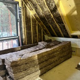 Michał Wieczorek DOMY DREWNIANE ,SZKIELETOWE. OBSŁUGA KOMPLEKSOWA - Poddasze w trakcie izolacji, widoczne krokwie wypełnione wełną mineralną oraz stos wełny przygotowany do montażu, nóż na wierzchu stosu, okno dachowe z widokiem na las.