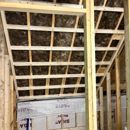 Michał Wieczorek DOMY DREWNIANE ,SZKIELETOWE. OBSŁUGA KOMPLEKSOWA - Wnętrze poddasza w trakcie ocieplania: widoczne drewniane belki konstrukcyjne, brązowa wełna mineralna między krokwiami oraz folia paroizolacyjna na ścianie kolankowej.