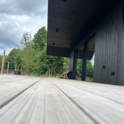 Michał Wieczorek DOMY DREWNIANE ,SZKIELETOWE. OBSŁUGA KOMPLEKSOWA - Nowoczesny dom z czarną elewacją w Trąbkach Wielkich, widok z poziomu drewnianego tarasu, na którym stoi rozłożone krzesło turystyczne i skrzynki z narzędziami.