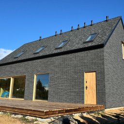Michał Wieczorek DOMY DREWNIANE ,SZKIELETOWE. OBSŁUGA KOMPLEKSOWA - Nowoczesny dom z czarnej cegły z drewnianym tarasem, okna z jasnymi ramami, dach pokryty szarą dachówką, widoczne kominy i okna dachowe, błękitne niebo w tle.