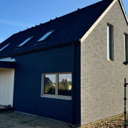 Michał Wieczorek DOMY DREWNIANE ,SZKIELETOWE. OBSŁUGA KOMPLEKSOWA - Nowoczesny dom z ciemną elewacją z cegły klinkierowej i drewnianym wejściem, widoczne rusztowanie i teren budowy.