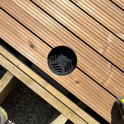 Michał Wieczorek DOMY DREWNIANE ,SZKIELETOWE. OBSŁUGA KOMPLEKSOWA - Drewniany taras z odpływem liniowym, widoczne elementy konstrukcyjne i podłoże z kamyków, fragment buta roboczego w rogu.